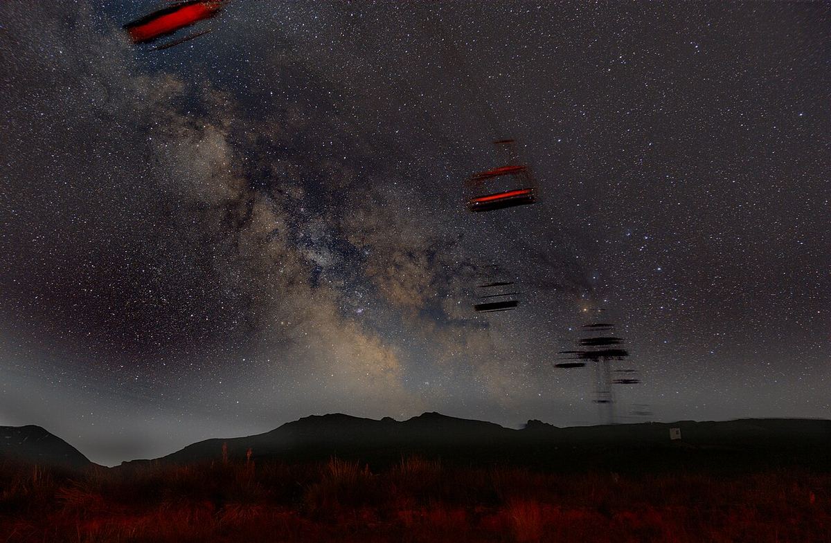 Miniatura: Via Láctea sobre montanhas escuras com cadeiras de um teleférico em primeiro plano