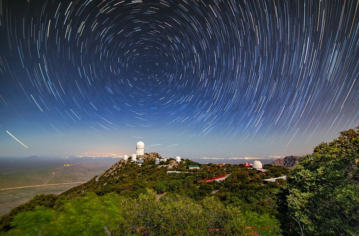 Rastos de estrelas sobre um observatório de montanha com o brilho de cidades no horizonte