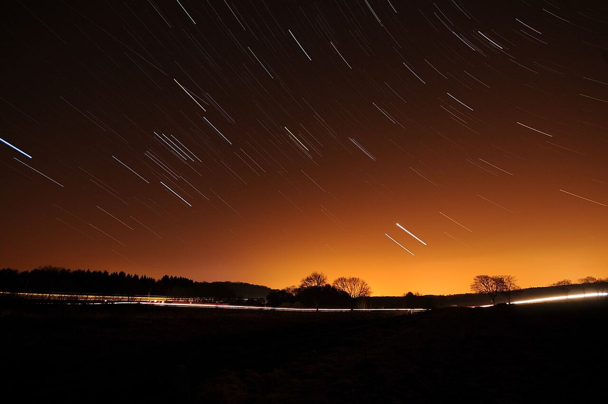 Miniatura: Rastos de estrelas sobre o horizonte com o brilho alaranjado de uma cidade