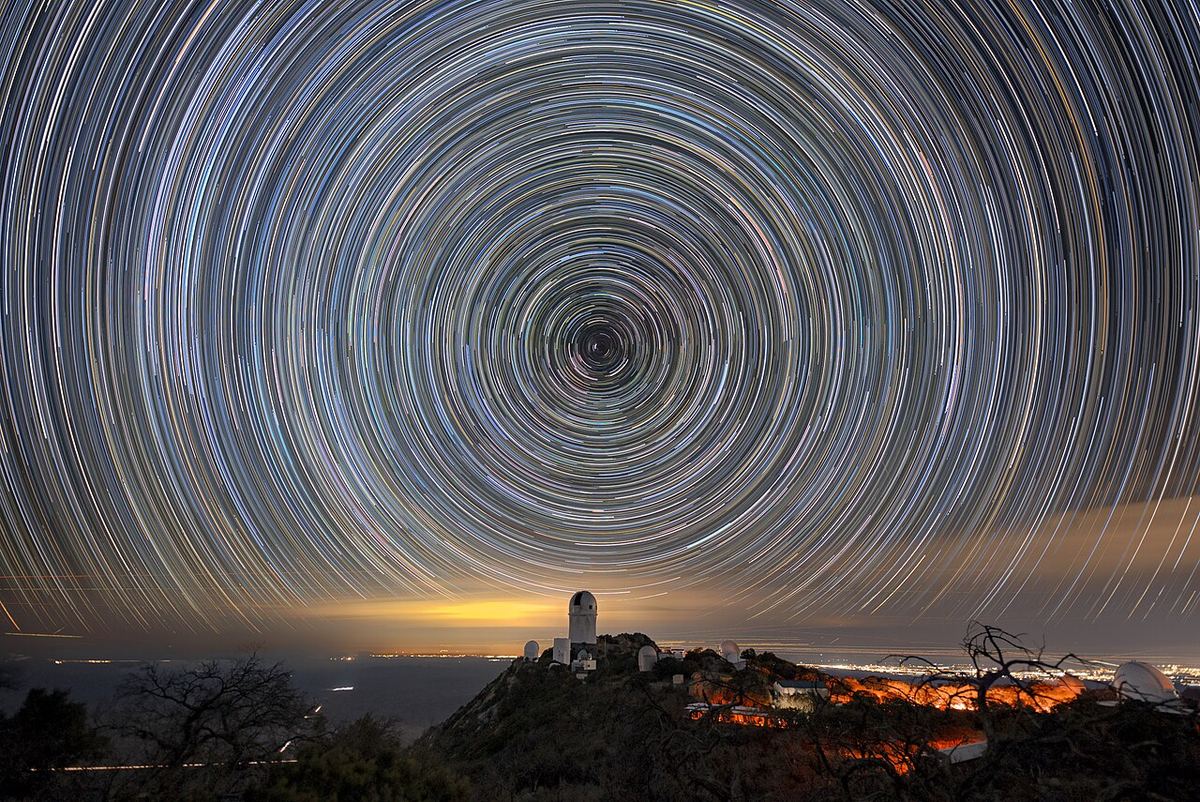Rastos de estrelas sobre as cúpulas de um observatório com o brilho de uma cidade no horizonte