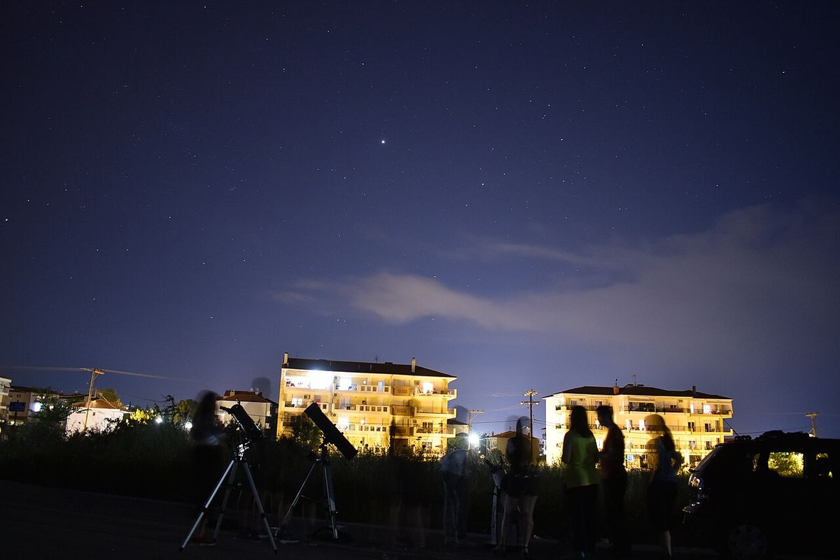 Miniatura: Telescópios montados num terreno ao anoitecer com edifícios ao fundo